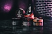 Various containers of tobacco products are stacked together on a dark surface with a dark purple and black background. The containers display different brands and flavors, with labels in multiple languages. A black hookah is partially visible in the background.
