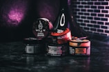 Various containers of tobacco products are stacked together on a dark surface with a dark purple and black background. The containers display different brands and flavors, with labels in multiple languages. A black hookah is partially visible in the background.