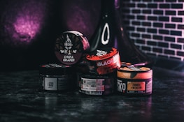 Various containers of tobacco products are stacked together on a dark surface with a dark purple and black background. The containers display different brands and flavors, with labels in multiple languages. A black hookah is partially visible in the background.
