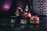 Various containers of tobacco products are stacked together on a dark surface with a dark purple and black background. The containers display different brands and flavors, with labels in multiple languages. A black hookah is partially visible in the background.
