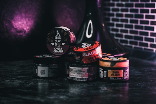 Various containers of tobacco products are stacked together on a dark surface with a dark purple and black background. The containers display different brands and flavors, with labels in multiple languages. A black hookah is partially visible in the background.