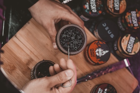 A person is preparing a hookah bowl with a dark, sticky substance. Several containers with different labels are scattered on a wooden board, possibly containing various flavors of shisha tobacco. The person is using a tool to pack the substance into the bowl.