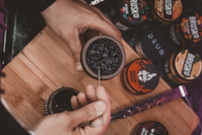 A person is preparing a hookah bowl with a dark, sticky substance. Several containers with different labels are scattered on a wooden board, possibly containing various flavors of shisha tobacco. The person is using a tool to pack the substance into the bowl.