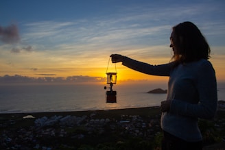 A person holds a lantern against the backdrop of a serene sunset by the sea. The silhouette of the individual is prominent, with the lantern illuminated by the sun's rays. The sky is filled with a blend of orange, yellow, and blue hues, creating a calm and picturesque view. Below, a coastal town and the ocean stretch into the distance.