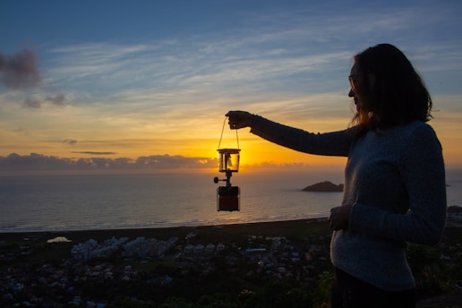 A person holds a lantern against the backdrop of a serene sunset by the sea. The silhouette of the individual is prominent, with the lantern illuminated by the sun's rays. The sky is filled with a blend of orange, yellow, and blue hues, creating a calm and picturesque view. Below, a coastal town and the ocean stretch into the distance.