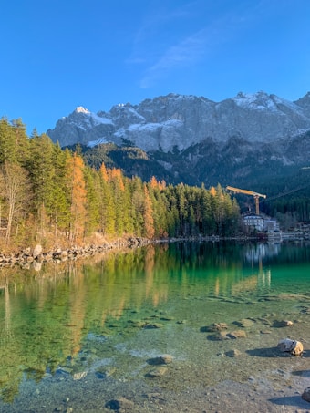Clear lake with a rocky shoreline reflecting surrounding trees and a distant mountain range, partially covered in snow under a bright blue sky. A construction crane and a building are visible near the forest edge.