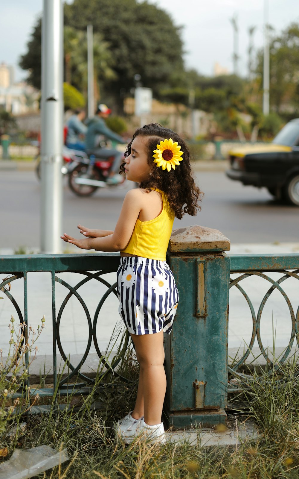 a little girl standing next to a fence with a sunflower in her hair