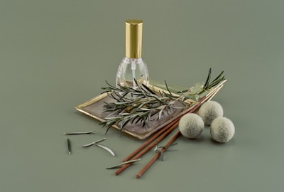 A small clear glass bottle with a gold cap is placed on a rectangular dish, surrounded by sprigs of rosemary. Next to it are two brown incense sticks and three round, fuzzy green balls, all set against a muted green background.