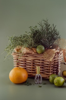 A woven basket filled with fresh herbs such as rosemary and thyme, accompanied by citrus fruits including an orange and limes on a green background. A small perfume bottle with a golden cap is situated in front of the basket.