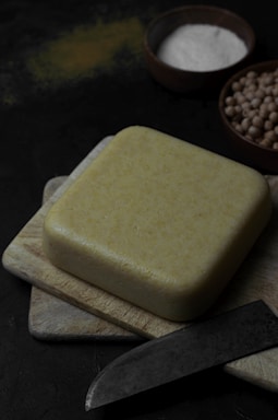A large block of yellow cheese sits on a wooden cutting board, accompanied by a dark-handled knife. Surrounding the cheese are bowls, one containing white powder (likely salt) and another with chickpeas. The overall setting is dimly lit with a dark background, highlighting the rustic and organic aspects of the ingredients.