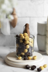 A glass jar filled with yellow and dark brown round cereal pieces is placed on a circular ceramic stand. The background features a kitchen setting with wooden utensils and ceramic cups, conveying a cozy and inviting atmosphere.