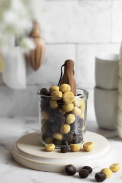 A glass jar filled with yellow and dark brown round cereal pieces is placed on a circular ceramic stand. The background features a kitchen setting with wooden utensils and ceramic cups, conveying a cozy and inviting atmosphere.
