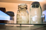 Three vintage glass jars are displayed on a shelf, with visible embossed lettering indicating brands such as Atlas and Mason. The jars have metal lids and appear to be well-preserved, contributing to a nostalgic and historical atmosphere.