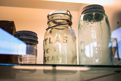 Three vintage glass jars are displayed on a shelf, with visible embossed lettering indicating brands such as Atlas and Mason. The jars have metal lids and appear to be well-preserved, contributing to a nostalgic and historical atmosphere.