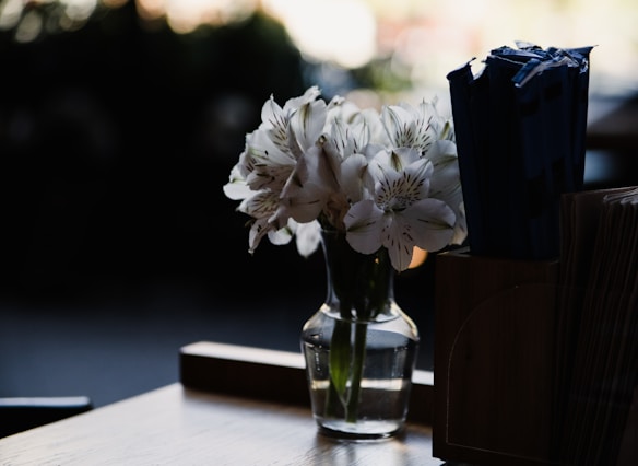 A small glass vase holds a bouquet of white lilies placed on a wooden table. The surrounding area is dimly lit, with soft natural light highlighting the delicate petals of the flowers. To the right, there is a container with numerous dark-colored sticks or packaged items. The background is out of focus, creating a serene and intimate atmosphere.