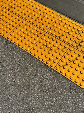 A textured yellow tactile paving with raised domes is set against a rough asphalt surface. The contrast between the bright yellow tiles and the dark gray ground is prominent, highlighting the functionality for visually impaired pedestrians.