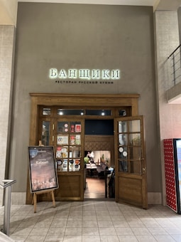 The entrance to a restaurant features a wooden doorway with glass panels. Above the door, there is a sign with Cyrillic text. A poster stand is positioned to the left of the entrance with information and graphics. The interior of the restaurant is visible through the door, revealing cozy seating and a warm, inviting atmosphere.