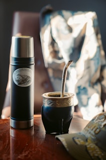A black thermos with a silver lid and logo stands on a wooden table next to a traditional mate cup filled with a metal straw. A camo-patterned jacket is draped over a chair in the background, and a cap is partially visible on the table.