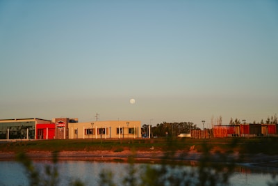 A commercial area with several buildings situated near a body of water under a clear sky with a visible moon. The buildings feature modern architecture with flat roofs and large windows, and the scene is bathed in warm, late-afternoon light. Sparse vegetation is visible in the foreground, and trees can be seen in the background.