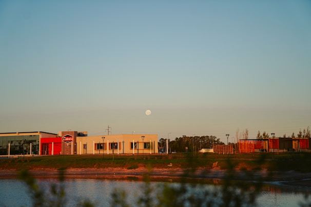 A commercial area with several buildings situated near a body of water under a clear sky with a visible moon. The buildings feature modern architecture with flat roofs and large windows, and the scene is bathed in warm, late-afternoon light. Sparse vegetation is visible in the foreground, and trees can be seen in the background.