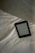 A digital e-reader device is placed on a white, wrinkled fabric surface, possibly a bed. Part of a grey fabric, which could be a blanket or a pillow, is visible in the top right corner. The e-reader screen displays text from an electronic book or document.