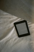 A digital e-reader device is placed on a white, wrinkled fabric surface, possibly a bed. Part of a grey fabric, which could be a blanket or a pillow, is visible in the top right corner. The e-reader screen displays text from an electronic book or document.
