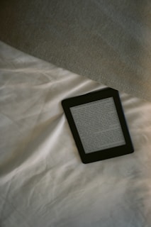 A digital e-reader device is placed on a white, wrinkled fabric surface, possibly a bed. Part of a grey fabric, which could be a blanket or a pillow, is visible in the top right corner. The e-reader screen displays text from an electronic book or document.