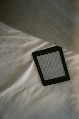 A digital e-reader device is placed on a white, wrinkled fabric surface, possibly a bed. Part of a grey fabric, which could be a blanket or a pillow, is visible in the top right corner. The e-reader screen displays text from an electronic book or document.