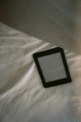 A digital e-reader device is placed on a white, wrinkled fabric surface, possibly a bed. Part of a grey fabric, which could be a blanket or a pillow, is visible in the top right corner. The e-reader screen displays text from an electronic book or document.