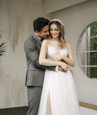 A couple embraces lovingly indoors, with the man wearing a gray suit and the woman in a white bridal gown. The woman wears a beaded headband and necklace while smiling and looking down. The interior background includes a round window and some greenery.