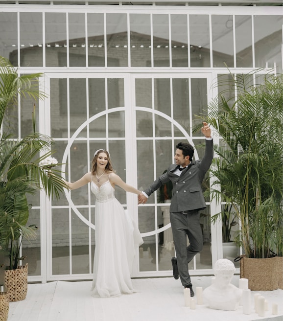 The couple holding hands, framed by burgundy and white floral arrangements with greenery accents.