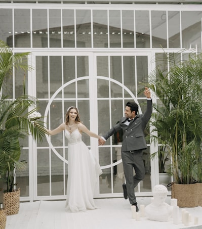 A couple joyfully holds hands, dressed in wedding attire inside a bright, plant-filled setting. The bride is in a flowing white gown and the groom in a sharp gray suit, both exuding happiness. They are surrounded by potted green plants, white candles, and a classic sculpture bust in a spacious, airy room with white grid windows.