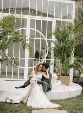 A couple sits closely together in a loving embrace, the woman wearing a white wedding dress and holding a bouquet of flowers, while the man is dressed in a gray suit. They are on a white, circular platform surrounded by green plants and white candles, set against a backdrop of large windows with a geometric design.