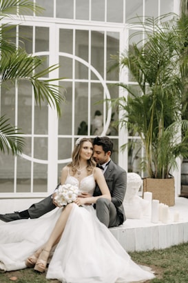 A couple dressed in formal wedding attire is sitting closely together. The bride is wearing a white gown and holding a bouquet of flowers, while the groom is in a dark suit. They are seated on a white platform surrounded by green plants and candles. Behind them is a large window with decorative white framing.