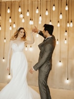 A couple dances gracefully, illuminated by a backdrop of hanging Edison bulbs. The woman wears an elegant bridal gown while the man is dressed in a tailored suit.