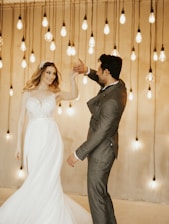 A couple dances gracefully, illuminated by a backdrop of hanging Edison bulbs. The woman wears an elegant bridal gown while the man is dressed in a tailored suit.