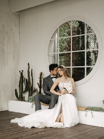 A couple is sitting close together in an intimate pose. The woman is wearing a long white wedding dress and holding a bouquet of flowers. The man is dressed in a formal suit with a bowtie, gently leaning towards her with an affectionate gesture. They are seated on a wooden floor beside a white wall decorated with cactuses and a large circular window.