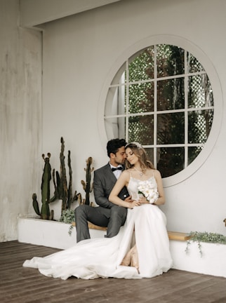 A couple is sitting close together in an intimate pose. The woman is wearing a long white wedding dress and holding a bouquet of flowers. The man is dressed in a formal suit with a bowtie, gently leaning towards her with an affectionate gesture. They are seated on a wooden floor beside a white wall decorated with cactuses and a large circular window.