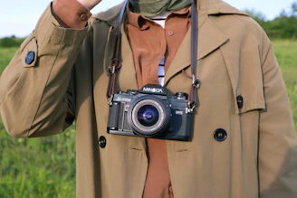 An outdoor shot of a person buttoning up a classic tan trench coat with a blurred park background.