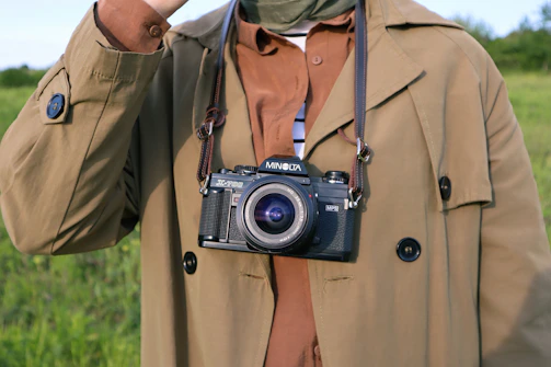 An outdoor shot of a person buttoning up a classic tan trench coat with a blurred park background.