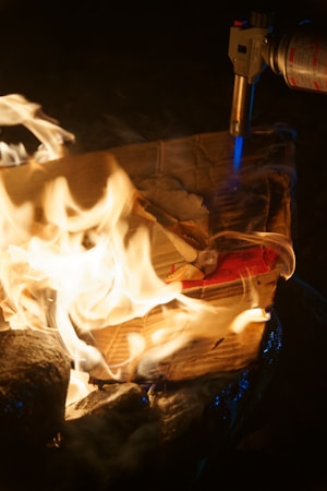 A cardboard box is burning with bright yellow flames, while a blowtorch emits a blue flame onto the box. The scene appears to be at night, indicated by the dark background. The fire is intense and engulfs parts of the cardboard, creating an interplay of light and shadow.