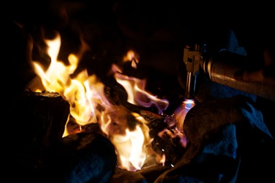 Flames rise among a collection of rocks in a dimly lit setting. A hand-held butane torch is being used, with its blue flame visible, adding to the intensity of the fire.