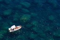 A small inflatable boat with a canopy floats on clear blue water, occupied by several people relaxing and sunbathing. The water reveals submerged rocks and gives a sense of tranquility.