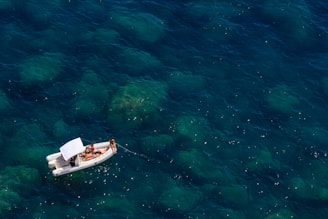 A small inflatable boat with a canopy floats on clear blue water, occupied by several people relaxing and sunbathing. The water reveals submerged rocks and gives a sense of tranquility.