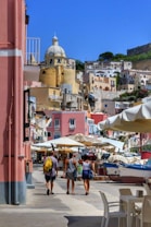 A vibrant coastal townscape featuring colorful buildings with a prominent dome structure. Several people walk along a street lined with market stalls and umbrellas, with a small boat visible nearby.