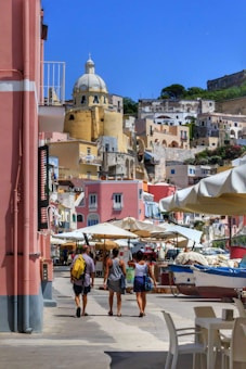 A vibrant coastal townscape featuring colorful buildings with a prominent dome structure. Several people walk along a street lined with market stalls and umbrellas, with a small boat visible nearby.