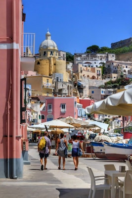 A vibrant coastal townscape featuring colorful buildings with a prominent dome structure. Several people walk along a street lined with market stalls and umbrellas, with a small boat visible nearby.