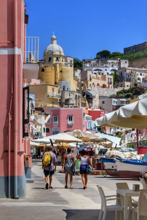 A vibrant coastal townscape featuring colorful buildings with a prominent dome structure. Several people walk along a street lined with market stalls and umbrellas, with a small boat visible nearby.