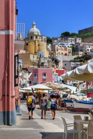 A vibrant coastal townscape featuring colorful buildings with a prominent dome structure. Several people walk along a street lined with market stalls and umbrellas, with a small boat visible nearby.