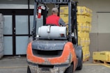 A man wearing a red vest operates a forklift in an industrial setting. The forklift appears weathered and is positioned near a large stack of yellow packaged materials labeled 'Premium'. There is a propane tank attached to the forklift, and a fire extinguisher is mounted nearby. The area is part of a warehouse or similar facility with large doors and additional pallets of materials in the background.